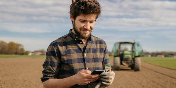 A farmer using a mobile phone in a field with a tractor in the background